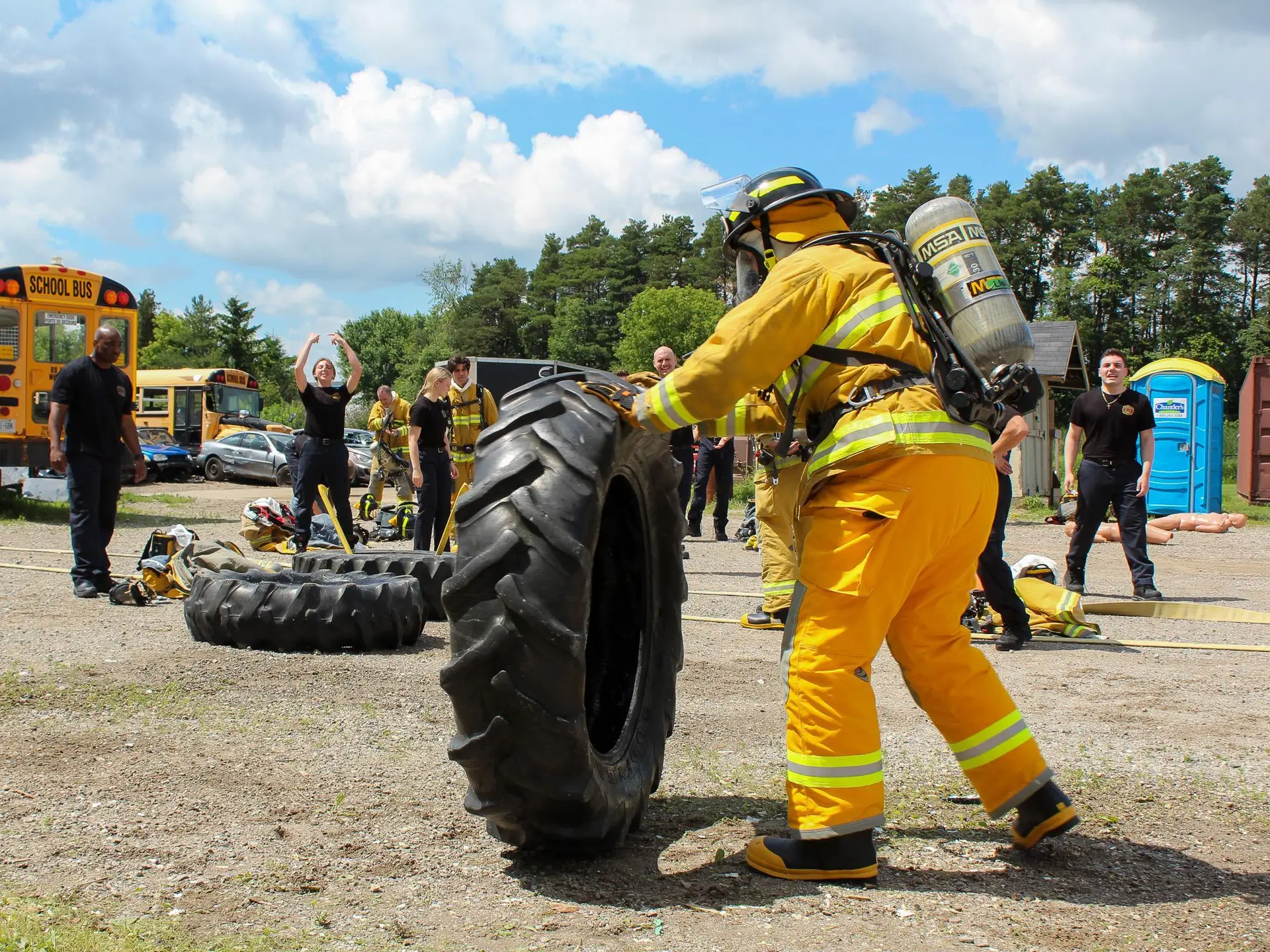 Firefighter recruits flipping large tires during outdoor training with a school bus in the background