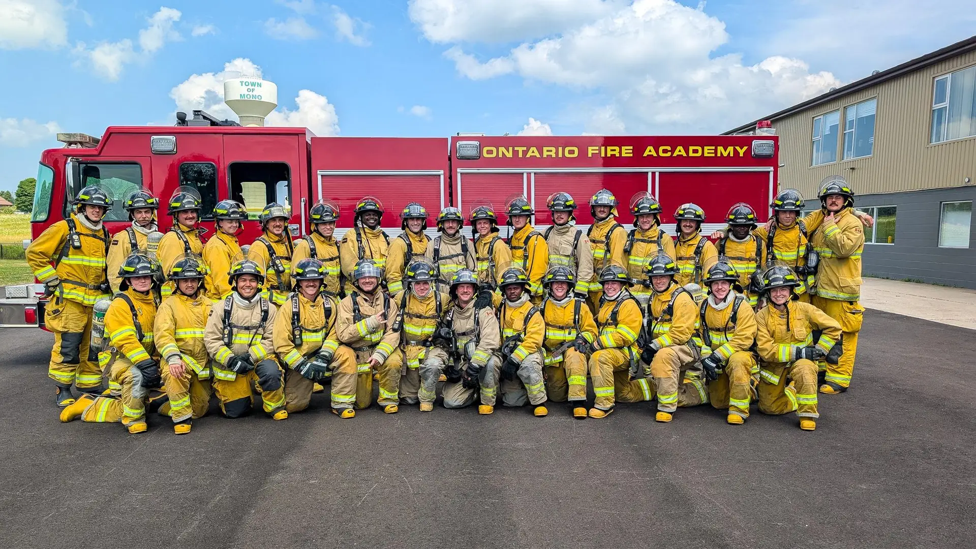 Group of firefighter recruits wearing full yellow turnout gear and helmets, posing in front of a red fire truck labeled "Ontario Fire Academy."