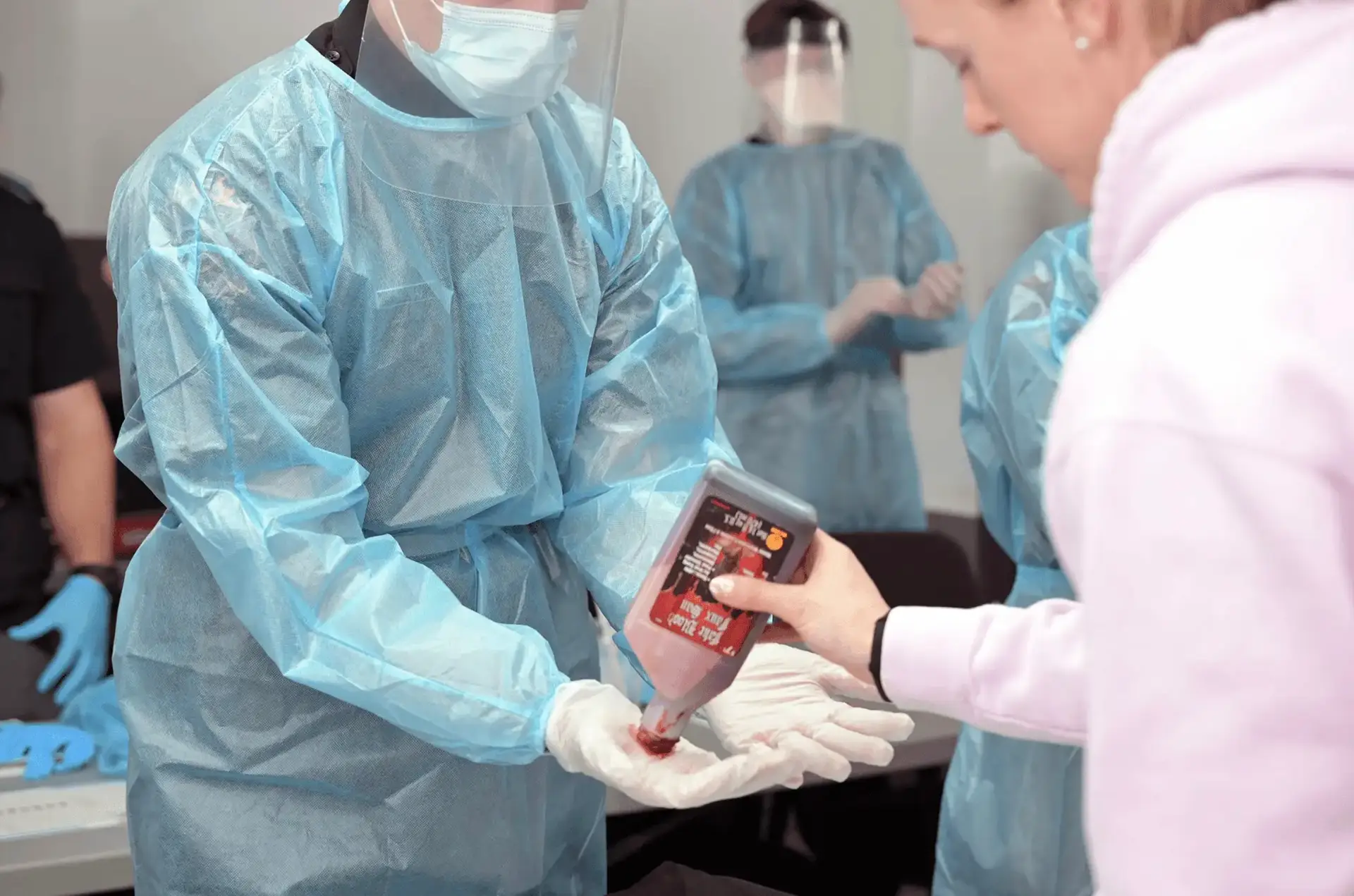 A first responder trainee in a blue protective gown, face shield, and gloves receives fake blood on their hands during a medical simulation.
